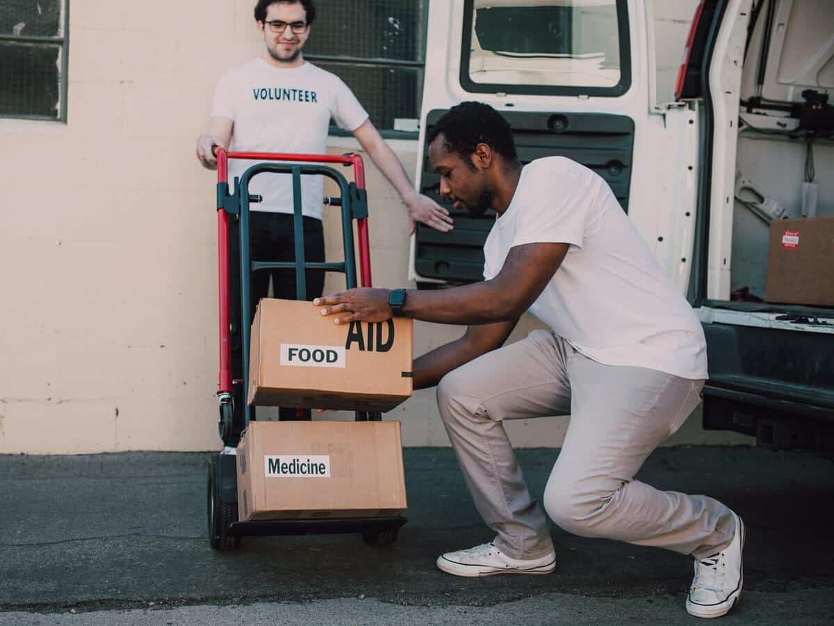 Two young men unload cardboard boxes from a white van onto a metal dolly.