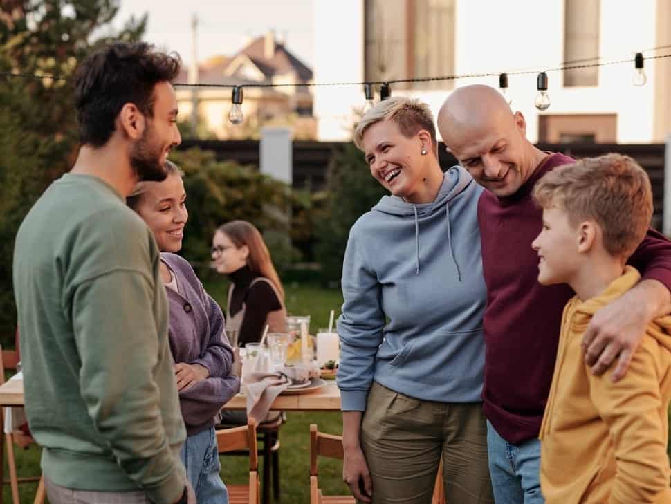 A group of adults and children are talking in the backyard of a home in front of a dinner table.