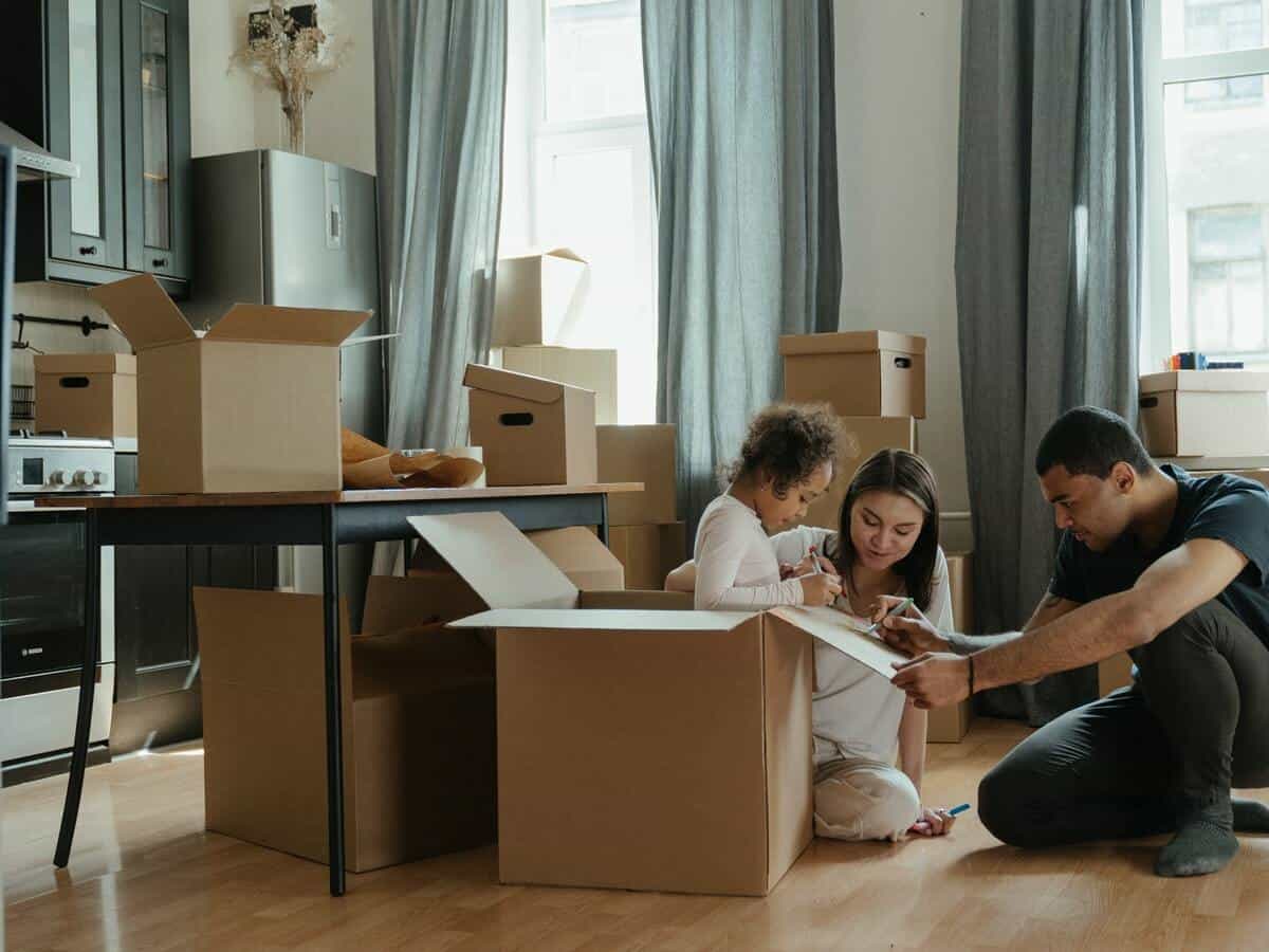 A young girl sits in a moving box as she and her parents decorate the exterior of the box while inside of their living room that’s surrounded by other moving boxes.