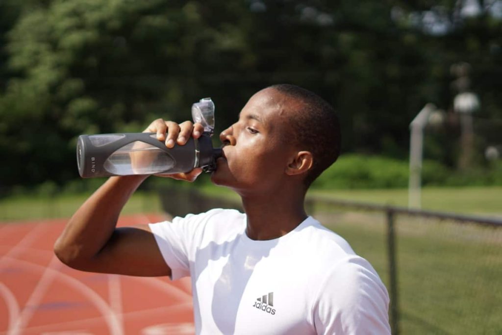 A young man standing on a track drinks water from a reusable bottle.