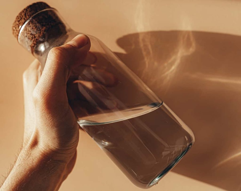 A closeup of a hand holding a glass water bottle in front of a light brown wall.