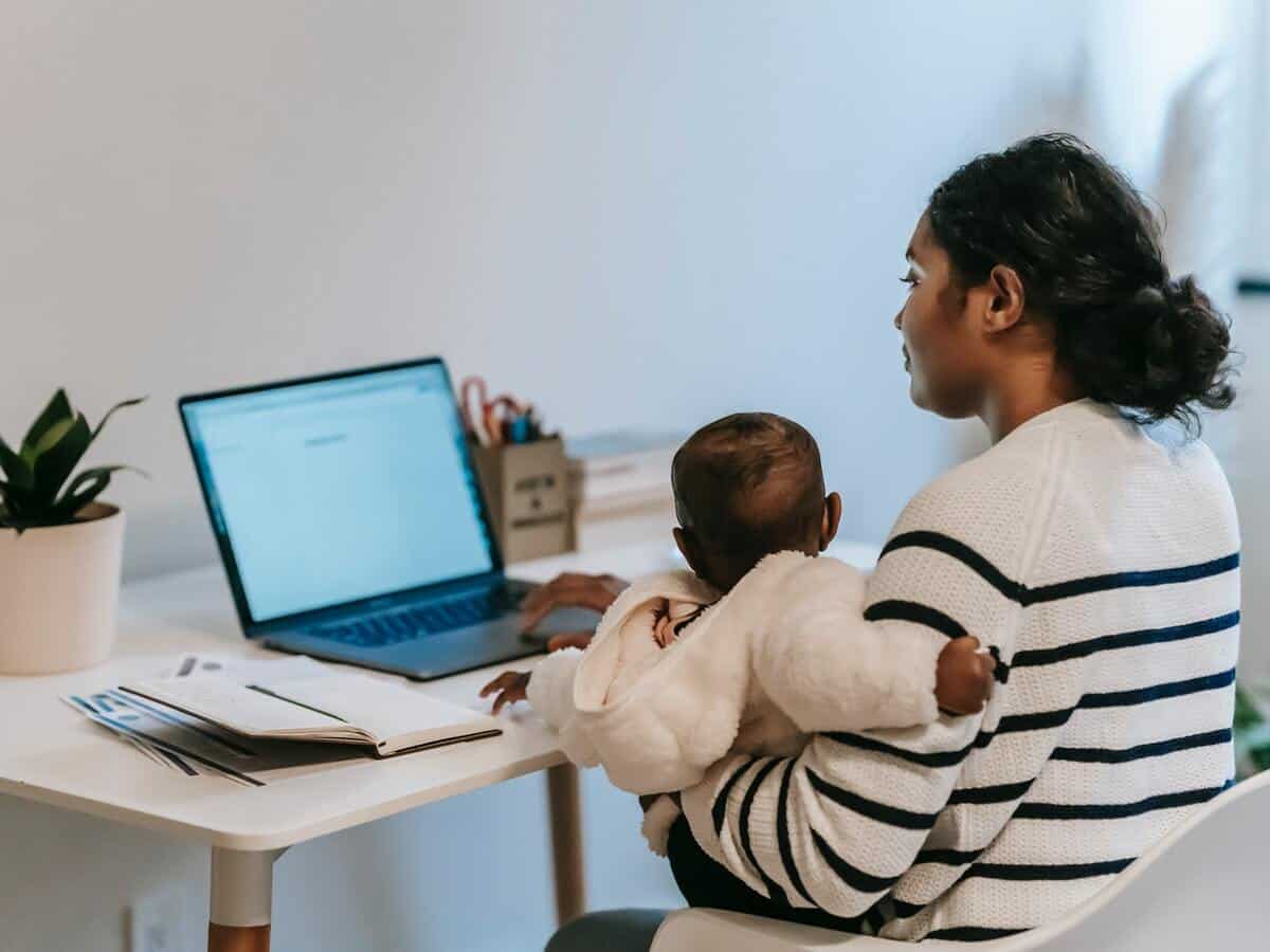 A woman is holding her baby while sitting at her desk and working on a laptop.