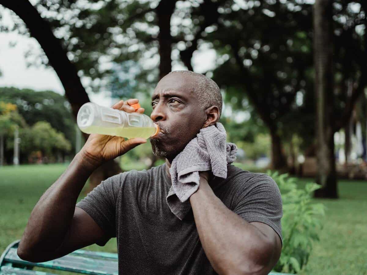 An older man with greying hair is drinking a sports drink while sitting outside in a park.