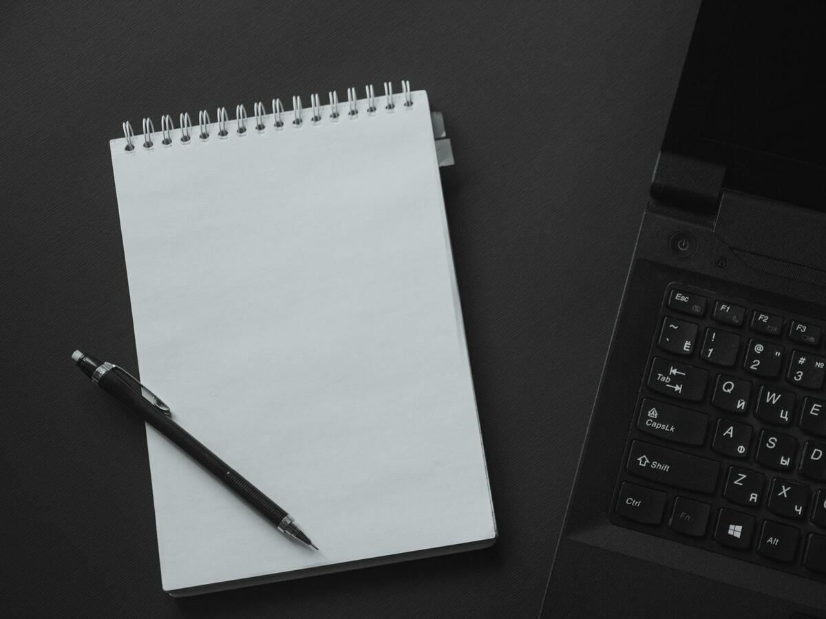 A white notebook with a black pen lying on top of it is next to a black laptop.