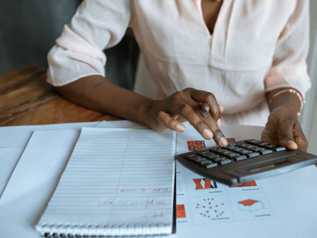 A woman is typing on a calculator while sitting at a table that's holding her notebook and other various documents.