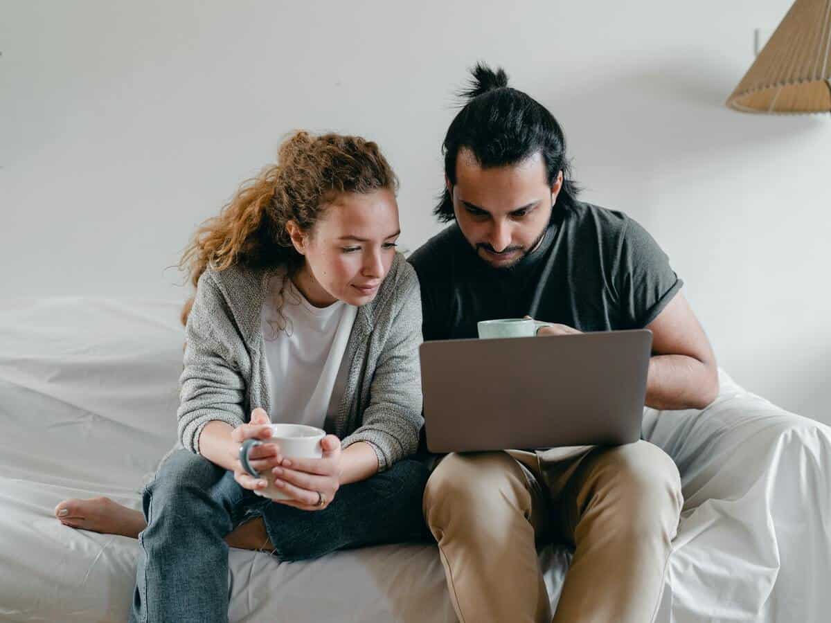 A young couple is looking at a laptop screen while each holding coffee cups.