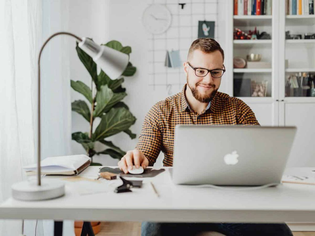 A young man wearing glasses smiles at his laptop while sitting at a desk inside his home.