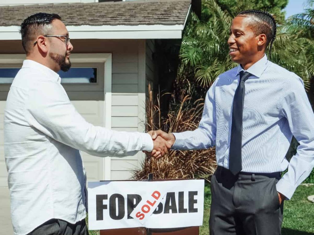 two men standing and shaking hands in front of a for sale and sold sign