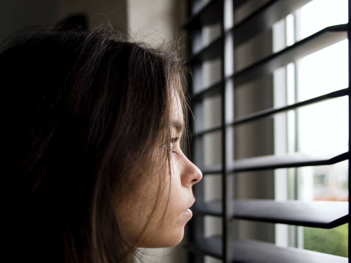 A woman is looking through the blinds of her window at home.