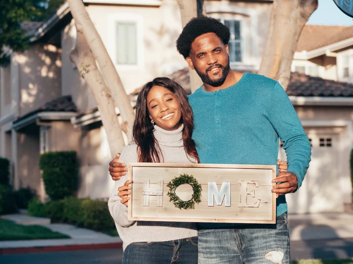 two new homebuyers standing in front of their new house holding a sign that says "home"