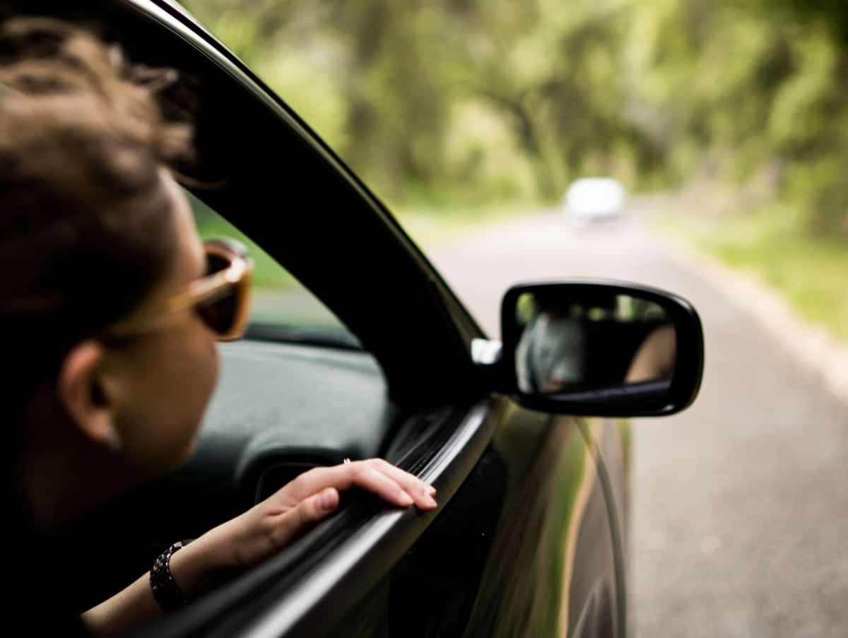 A woman wearing sunglasses has her head out the window while sitting in a car driving down a road that's surrounded by trees.
