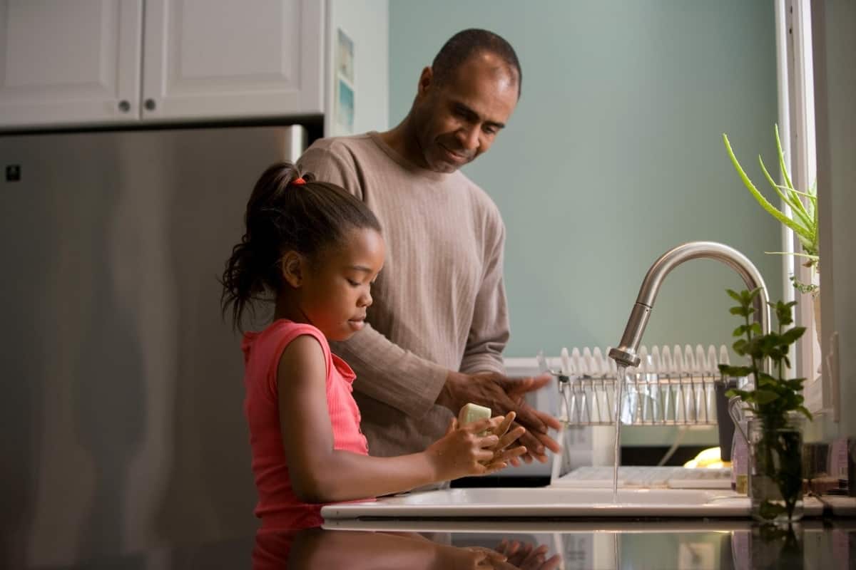A father teaches his young daughter how to wash her hands at the kitchen sink.