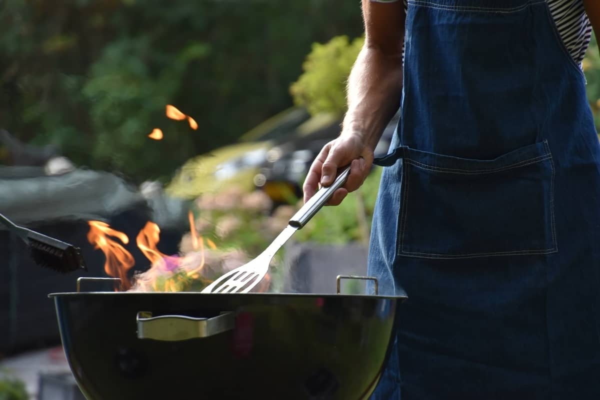 A man holds a spatula close to a black kettle grill outside.