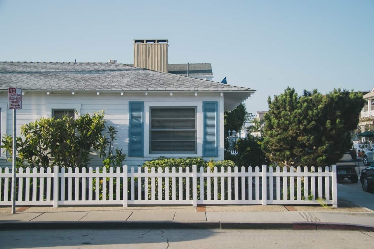 A white one-story house with blue shutters is on a street with parked cars.