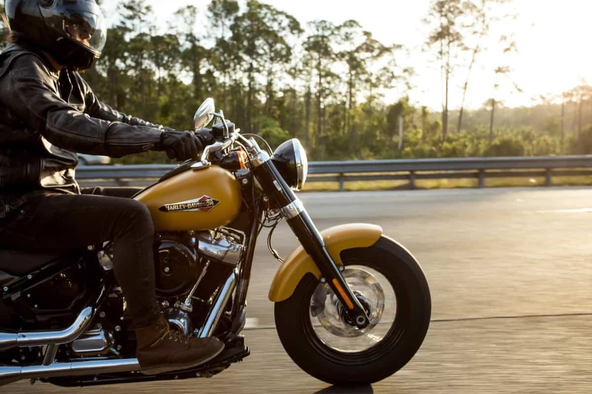 A man wearing a leather jacket, dark jeans, brown boots, and a helmet is riding his motorcycle down a road at sunset.