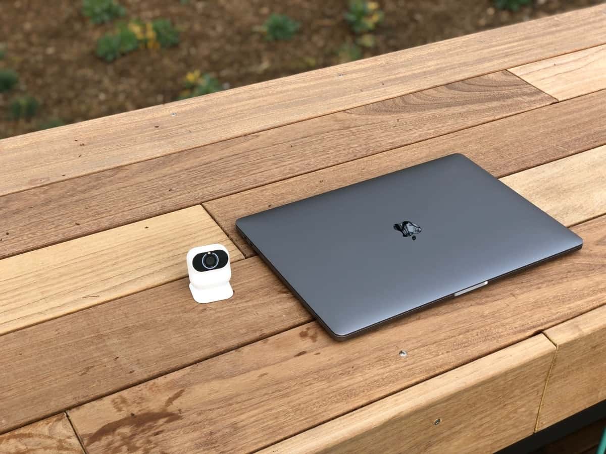 A white and black security camera sits next to a dark grey Macbook on a brown wooden bench.