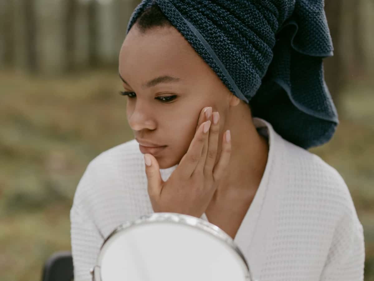 close up of a girl wearing a towel on her face, touching her face and looking in the mirror for acne or other blemishes