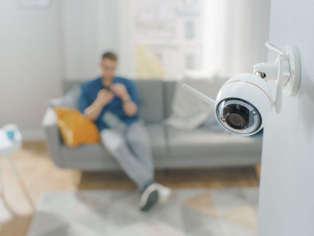 Close Up Object Shot of a Modern Wi-Fi Surveillance Camera with Two Antennas on a White Wall in a Cozy Apartment. Man is Sitting on a Sofa in the Background.