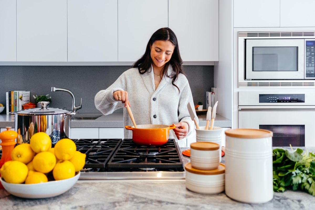 A woman with dark hair is stirring a dutch oven pot over a stove in a modern kitchen with fruit and vegetables on the counter.