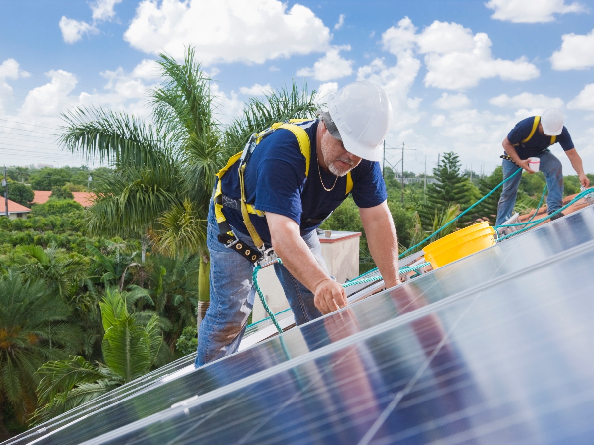 two men installing solar panels on a roof with palm trees and other homes in the background
